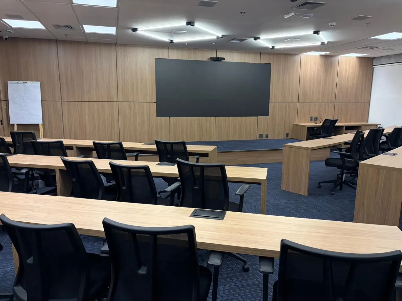 Sala de treinamento corporativa com painel de madeira, bancadas e iluminação linear